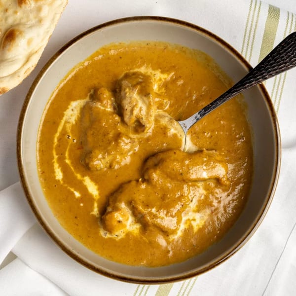 Overhead view of a bowl of creamy orange curry with chicken, a spoon, and naan bread.