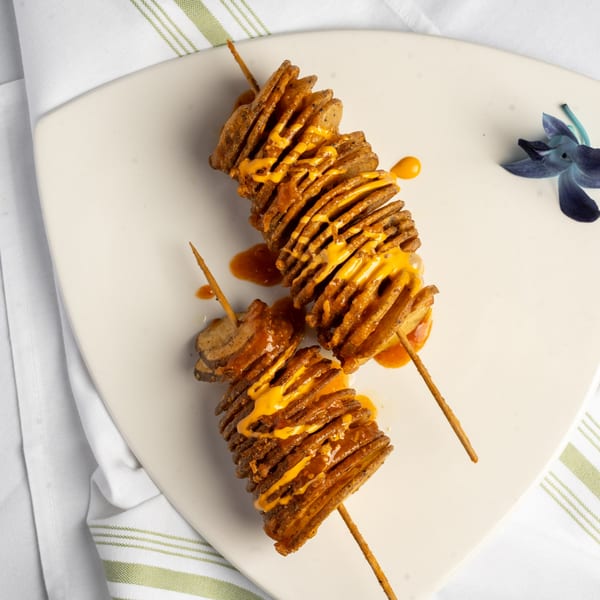 Two tornado potato skewers, drizzled with orange sauce, on a white plate with a decorative flower.