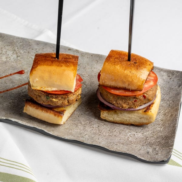Two mini burgers with patties, tomatoes, and onions on toasted buns, served on a gray plate.