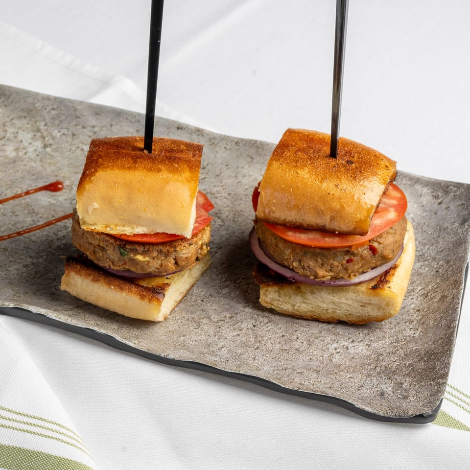 Two mini burgers with patties, tomatoes, and onions on toasted buns, served on a gray plate.