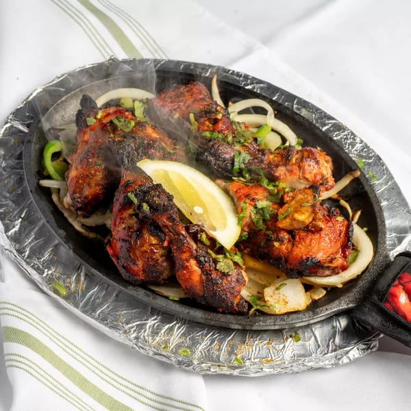 Sizzling tandoori chicken with onions, green peppers, cilantro, and a lemon wedge on a hot plate.